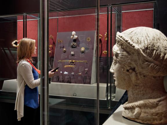 Visitor with multimedia handheld guide looking at ancient jewelry in display case, large sculpture in foreground