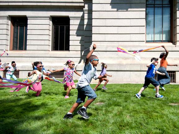 Kids in sunglasses running on Museum lawn, flying kites