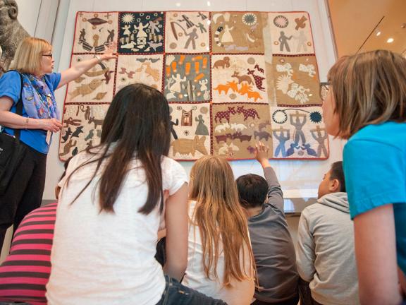Kids in school group sitting down, guide pointing at Harriet Powers quilt