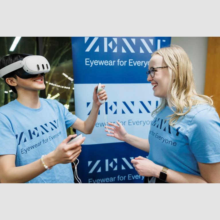 Person wearing virtual reality headset while holding controllers; another person watches, laughing