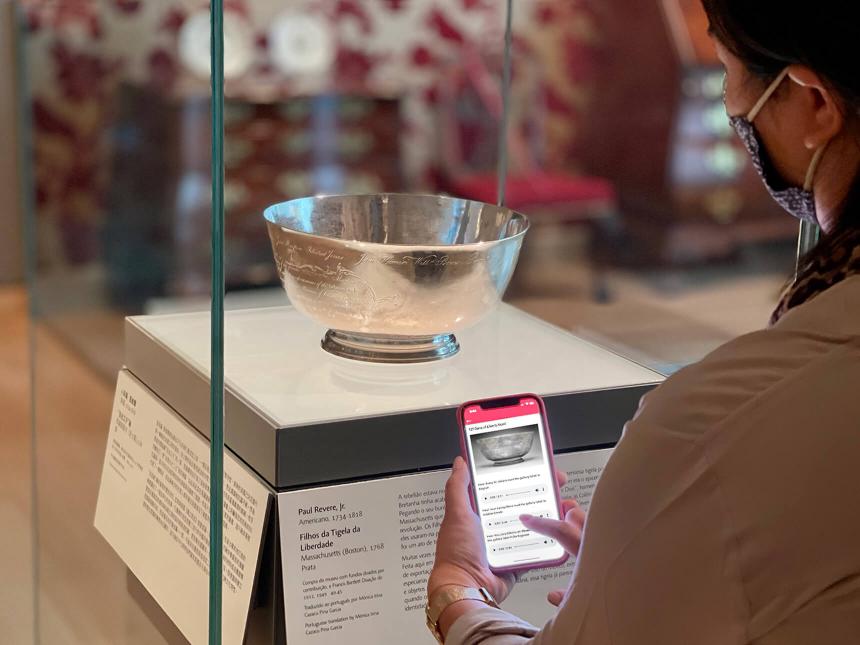 Visitor holding up smartphone with audio stop on screen, standing in front of silver Liberty Bowl in case