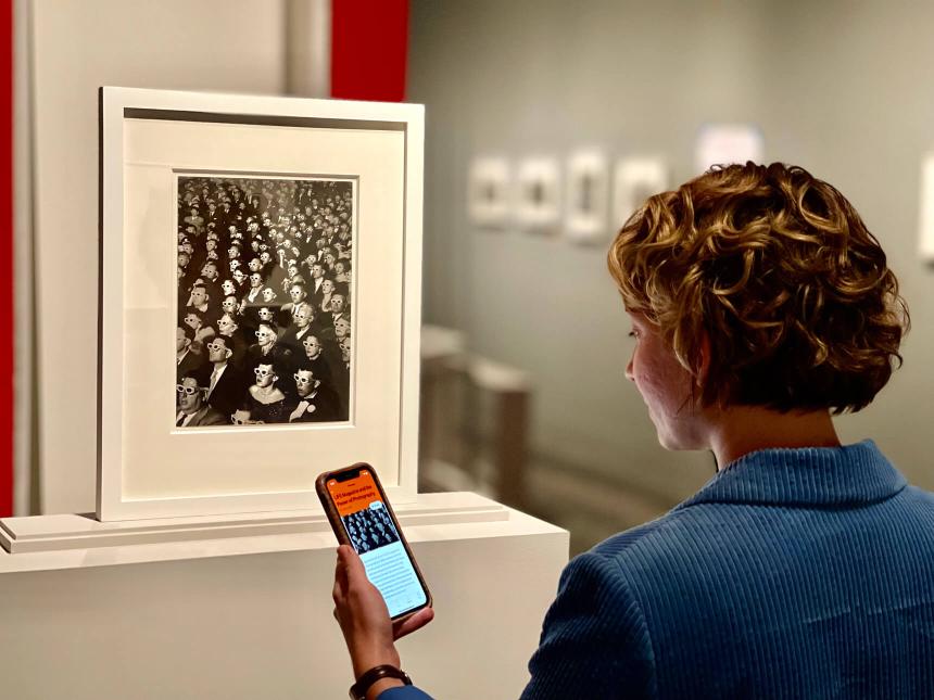 Visitor looks at smartphone loaded with MFA Mobile tour in front of framed black-and-white photograph of theater audience wearing 3-D glasses