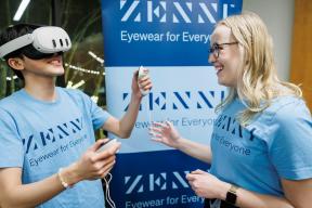 Person wearing virtual reality headset while holding controllers; another person watches, laughing