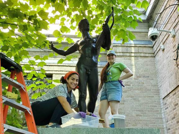 Two interns standing on stone pedestal with statue, working on cleaning sculpture