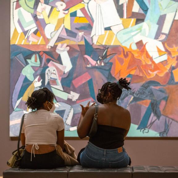 Two visitors sitting on bench in front of large abstract painting featuring many geometric shapes