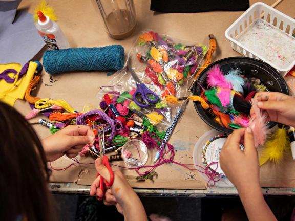 A craft table shown from above with colorful embroidery, feathers, and other art supplies on it