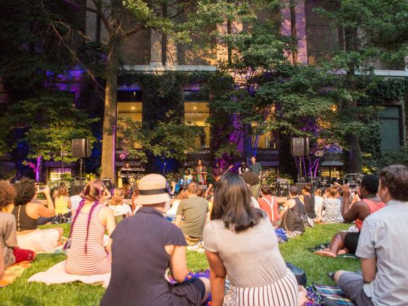 Concert-goers sit on blankets on the grass in a courtyard and watch musicians perform
