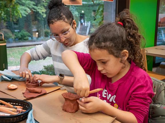 A woman and a child sit at a table and create pinch pots with clay.