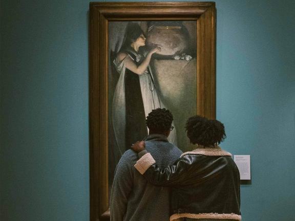 Two visitors looking up at large painting that depicts woman in sleeping gown seemingly asleep on bed, but with arms reaching out next to her as if embracing invisible person
