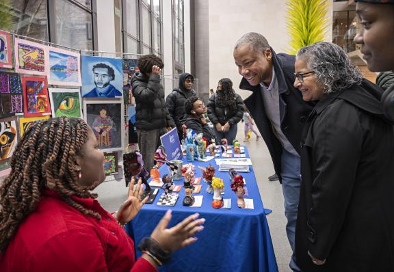 A small group of people gather around a young artist who sits next to a display of various artworks on a table and hanging behind her