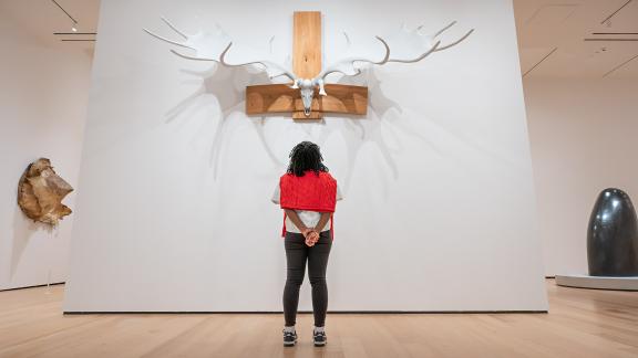 Visitor with red sweater around shoulders looks up at sculpture on wall made of large antlers mounted on animal skull