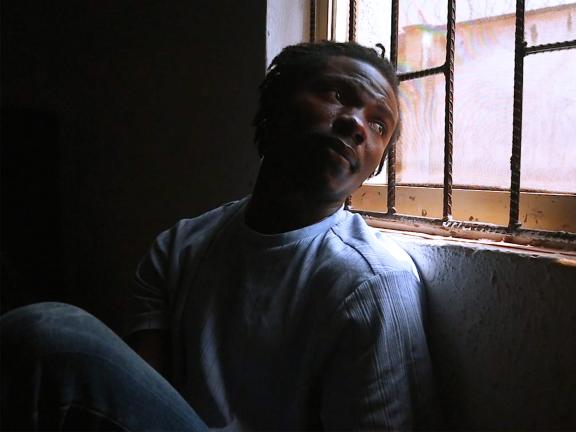 A man sits on the ground with his back to a wall, gazing over his shoulder out a window with bars on it