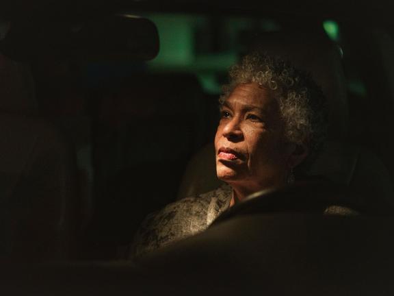 A woman sits behind the wheel of a car and is bathed in overhead light as she looks up and in the distance.