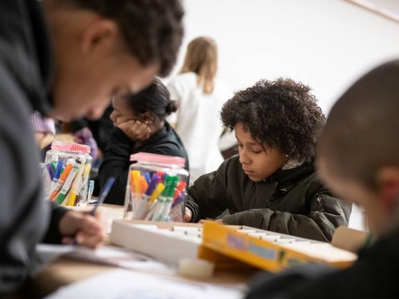 A group of young people participate in an art-making activity using colored markers on a communal table