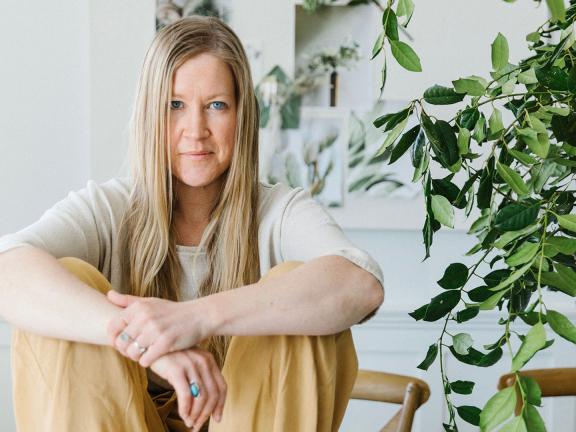 A woman sits on the ground with her arms on her knees next to a large house plant