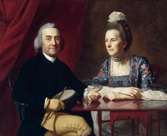Before a lush red fabric curtain, a man in a powdered wig sits at a mahogany table, next to a woman in a dress with an elaborate floral pattern and a small cap.