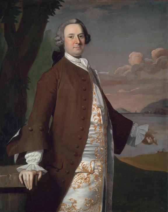 A colonial-era man in a powdered wig and brown coat stands before a coastal landscape wearing a satisfied grin.