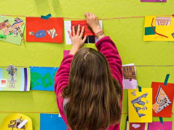 A child with brown hair hangs up an artwork on a brightly colored wall filled with other pieces of artwork