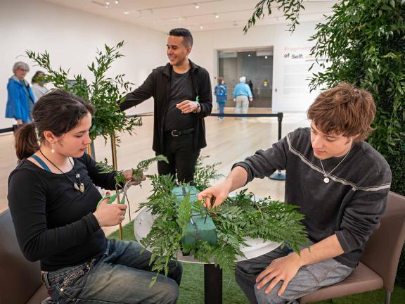Two young people in the foreground sit at a table working on preparing greenery and vegetation for a creative presentation