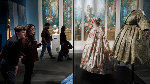 People stand in a dark blue gallery and look at ornately decorated floral dresses in glass cases.