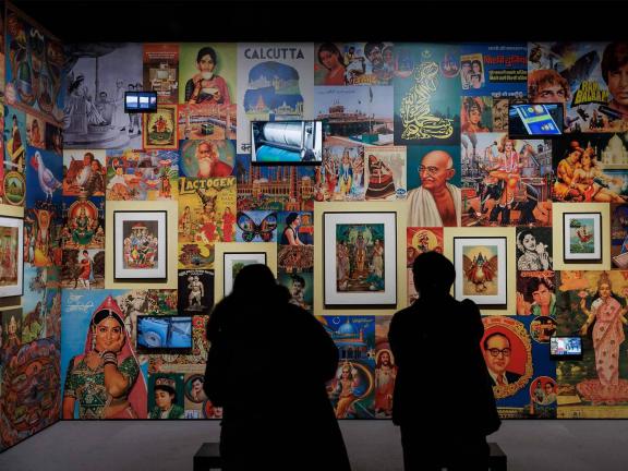 Two museum visitors appear as silhouettes in front of a colorful gallery wall of prints, ephemera, and video monitors