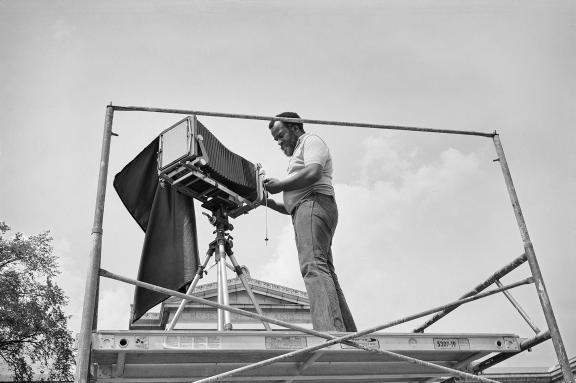 A man stands on a scaffold with a large-format camera.