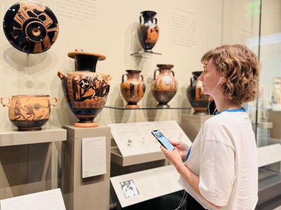 A woman with headphones engages on her phone while looking at ancient Greek objects in a gallery