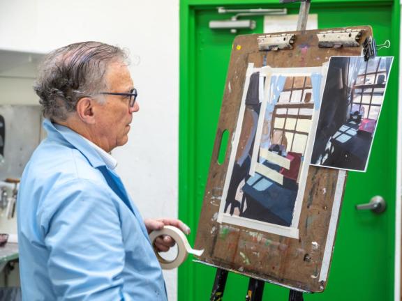 Adult male student in blue smock looks at painting on easel in classroom (SAC)