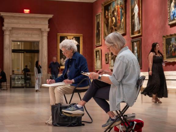 Two adult female students sit on stools and draw in sketchbook from the galleries (SAC)