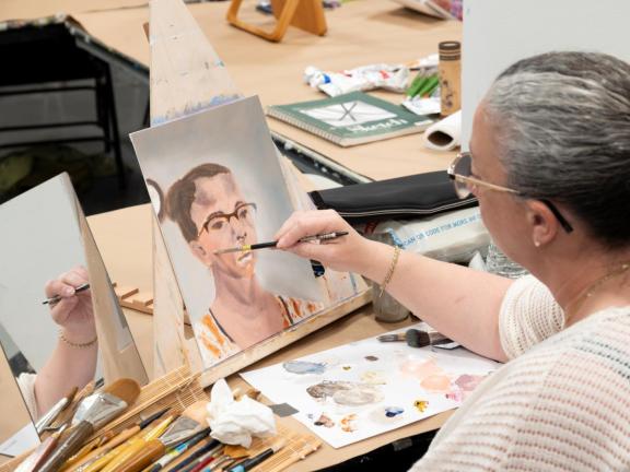 Adult female student paints self portrait on table top easel with mirror and paint brushes/palette (SAC)