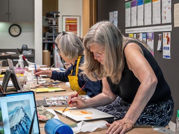 Two adult watercolor students paint at tables in classroom (SAC)