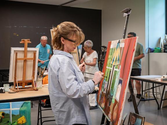 Adult female student paints on large canvas on easel in classroom with students in background (SAC)