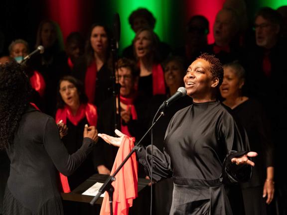 A woman stands at a microphone and sings with her arms raised.
