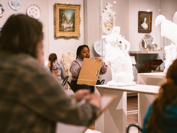 A person sits with a clipboard on their lap and draws while observing white porcelain bird sculptures in a brightly lit art gallery.