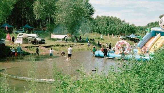 Photograph of people playing in a body of water with cars and campers and tents as well as inflatable water toys.