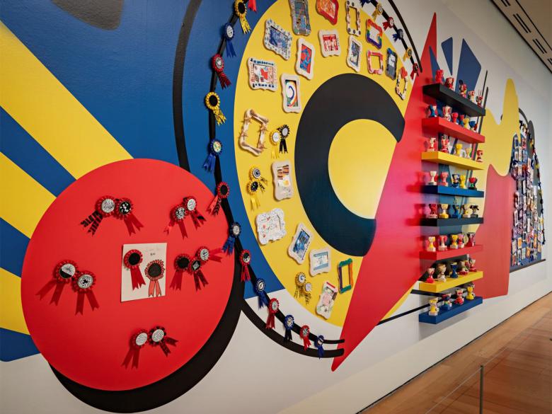 Red, yellow, and blue mural of various shapes with small sculptures and ribbons attached to the wall alongside it.
