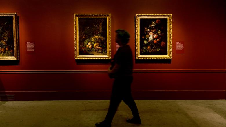 Silhouette of visitor walking past two still life paintings depicting flower arrangements in Rachel Ruysch exhibition
