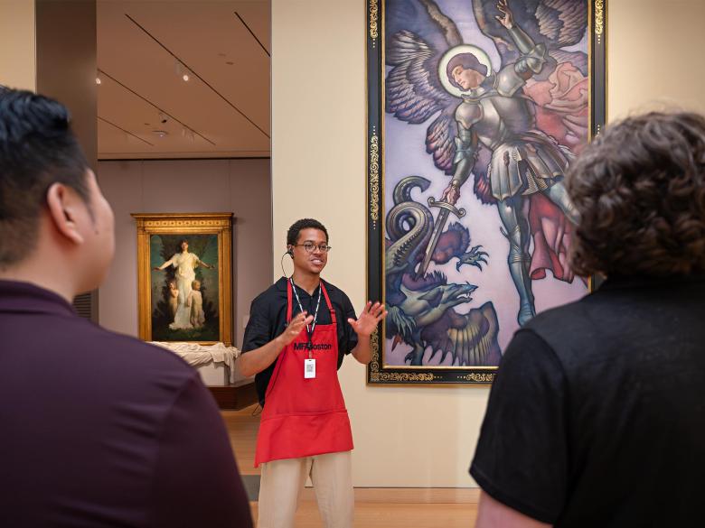 MFA Ambassador in red apron giving talk next to large painting depicting winged angel in armor pointing sword towards dragon; visitors standing in foreground
