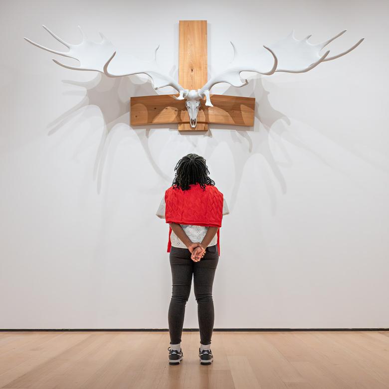 Visitor with red sweater around shoulders looks up at sculpture on wall made of large antlers mounted on animal skull