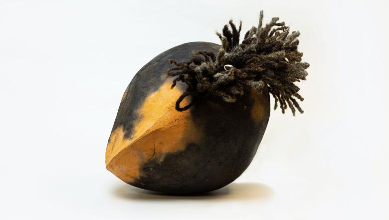 A yellow and black football-shaped object with locks of human hair sticking out of it leans on a white floor against a white backdrop.