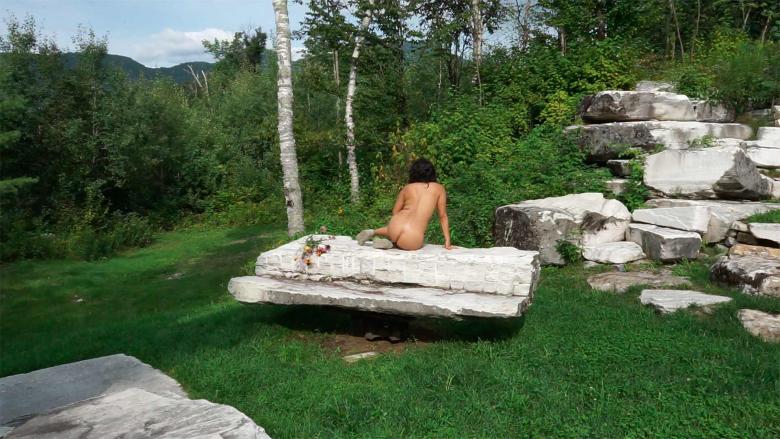 A nude figure sits on a white marble slab with their back turned to the frame, a green landscape surrounding them.