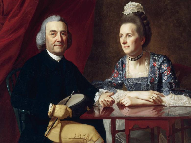 Before a lush red fabric curtain, a man in a powdered wig sits at a mahogany table, next to a woman in a dress with an elaborate floral pattern and a small cap.