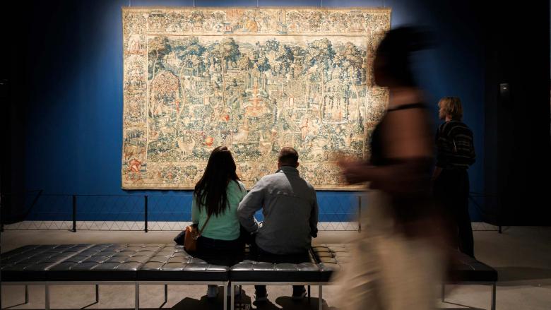 Two people sit on a bench and look at a large, ornately decorated tapestry hung on a dark blue wall.