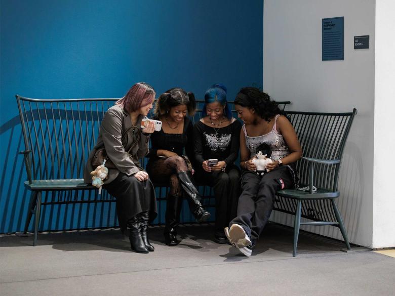 Four people sit on a dark green curved wooden bench in a corner of a room with blue and white walls.