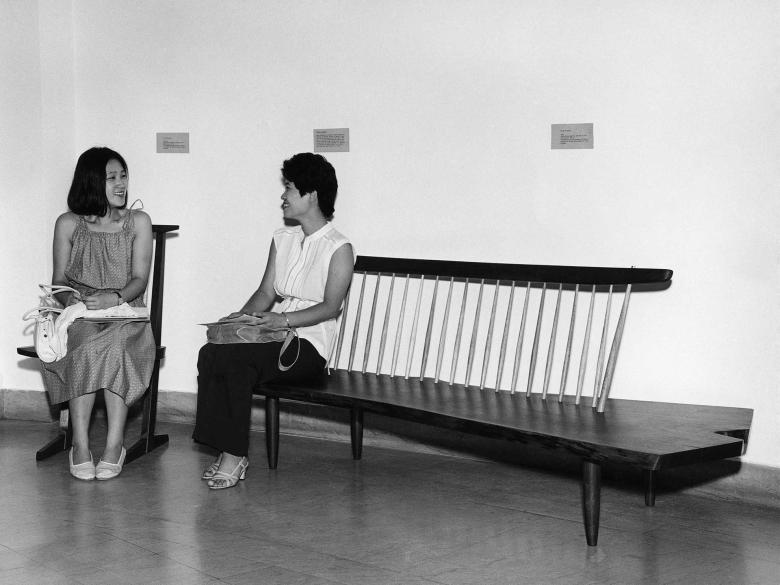 Two people sit on chairs in an empty space with white walls.