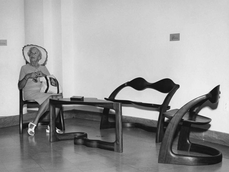 A woman wearing a wide-brimmed hat sits in a chair beside two empty chairs.