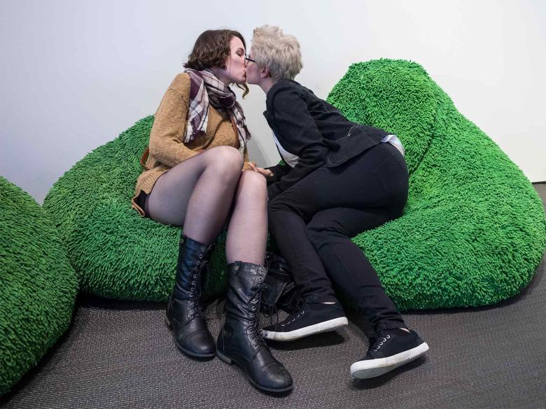 Two people sit in furry green bean bag chairs and kiss.