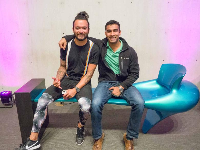 Two people sit on a futuristic teal bench, one with their arm around the other, and stare directly ahead smiling.