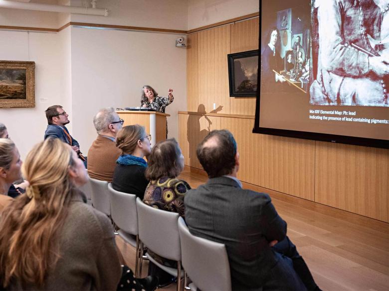 A person stands behind a podium and points to a presentation projected on a wall before a seated audience.