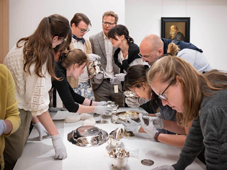 A group of people stand over a white table and inspect pieces of silver laid out across it.